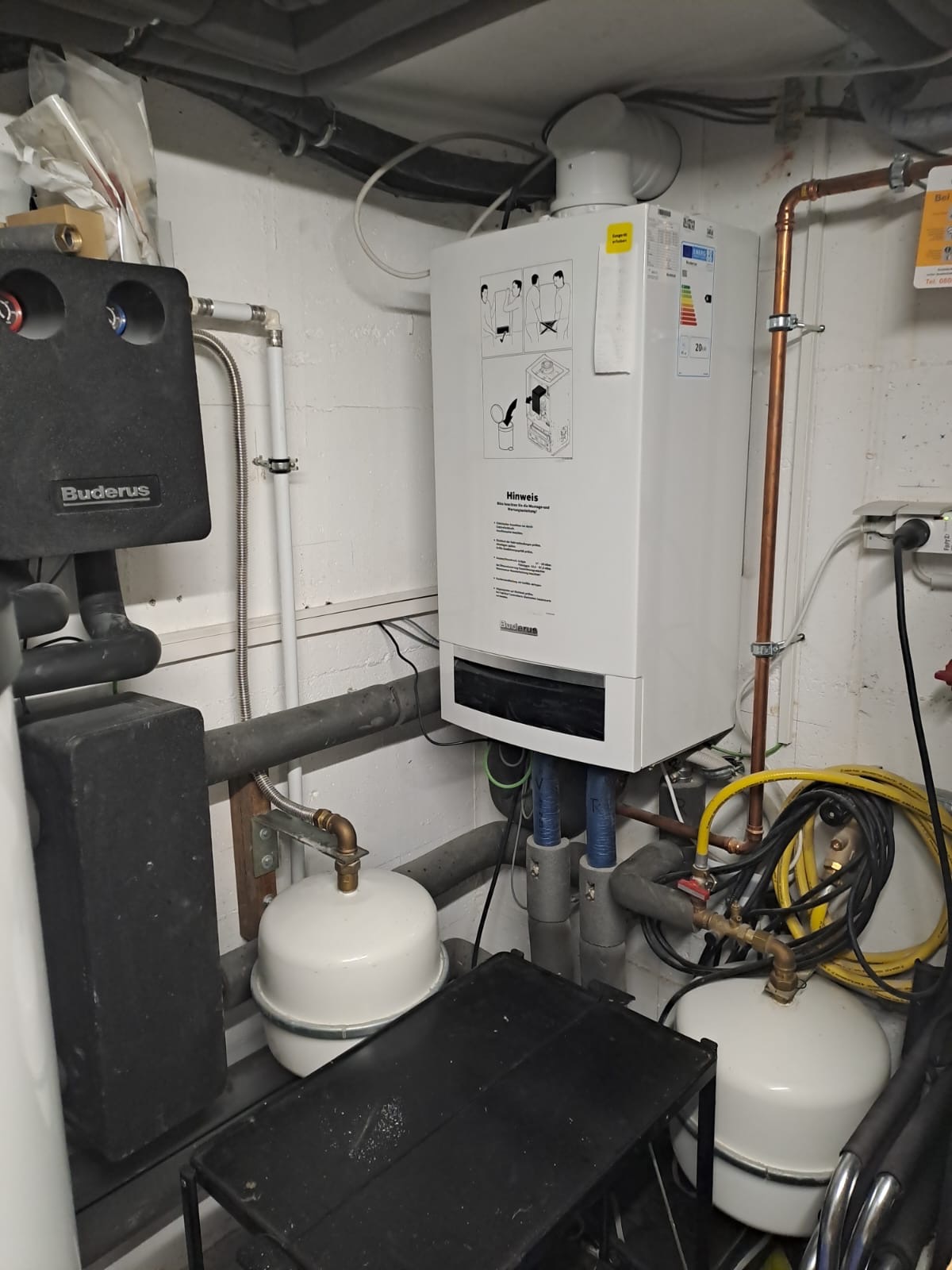 Nowoczesny, biały piec gazowy marki Buderus zamontowany na ścianie z widocznymi rurami miedzianymi i przewodami, w tle ściana z surowego betonu.