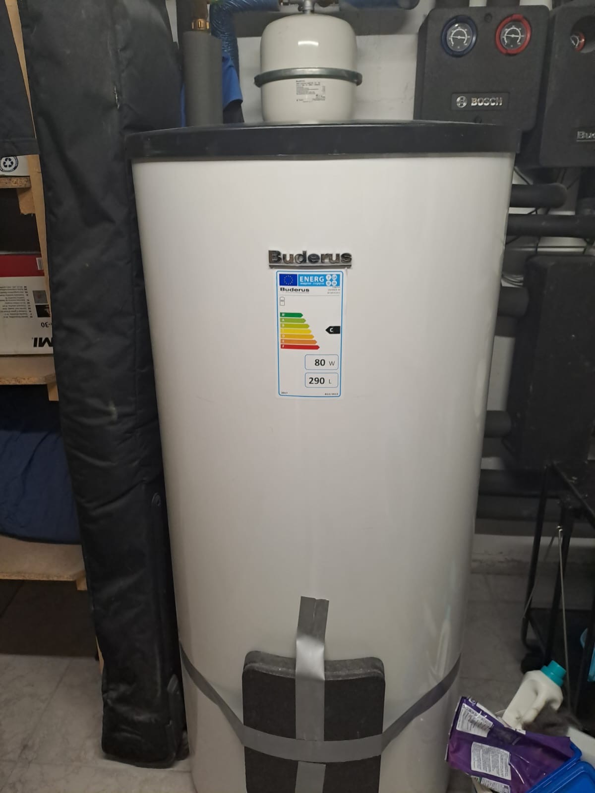 Biały zasobnik ciepłej wody użytkowej marki Buderus o pojemności 290 litrów, z widoczną etykietą energetyczną i zabezpieczeniem transportowym z szarej taśmy, stojący w kotłowni.