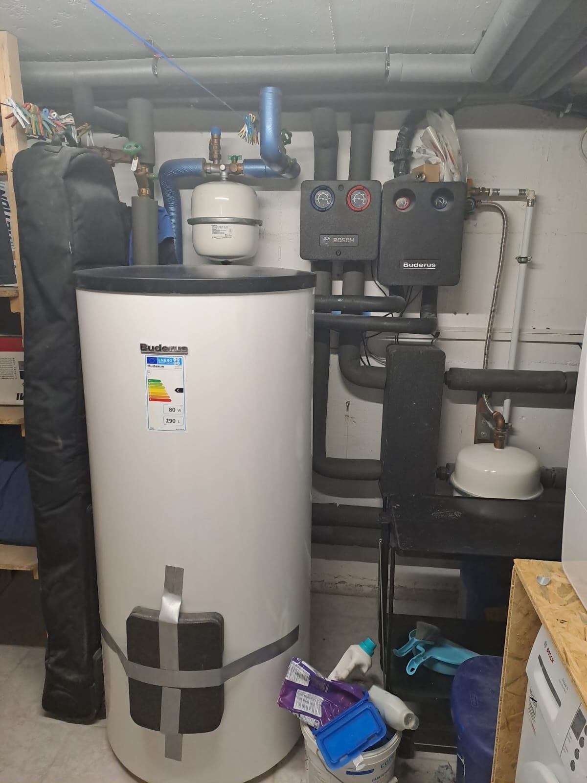 Biały bojler Buderus z etykietą energetyczną, podłączony do instalacji grzewczej z czarnymi rurami i dwoma panelami sterującymi Bosch i Buderus, widoczne również akcesoria hydrauliczne i pranie w tle.