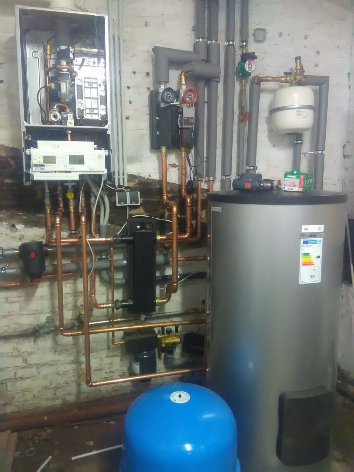 Nowoczesny piec centralnego ogrzewania z otwartą obudową, widoczne miedziane rury, szary zbiornik na wodę, niebieski hydrofor, narzędzia i akcesoria na górze zbiornika, surowa ściana z cegły w tle.