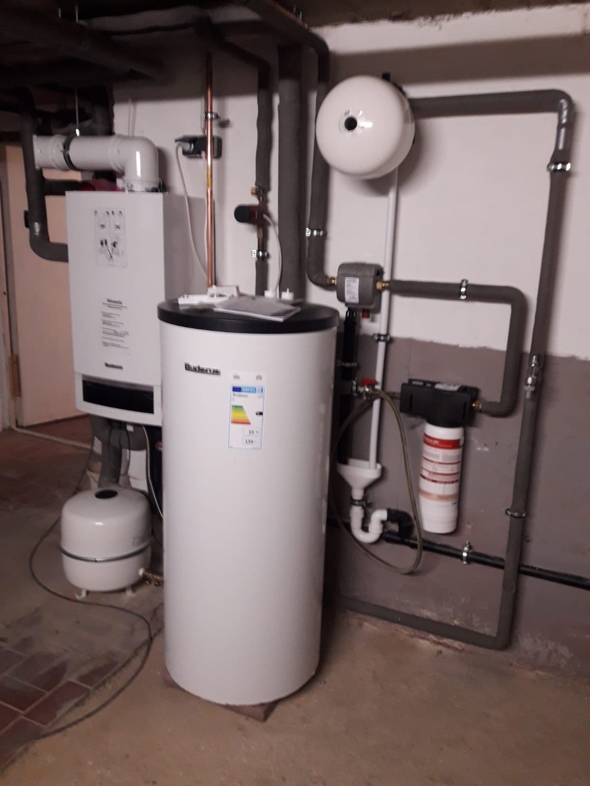 Biały zasobnik ciepłej wody użytkowej Buderus z czarnym blatem, obok wiszący kocioł gazowy, instalacja rur z izolacją termiczną, filtr wody, naczynie wzbiorcze, zamontowane w piwnicy.