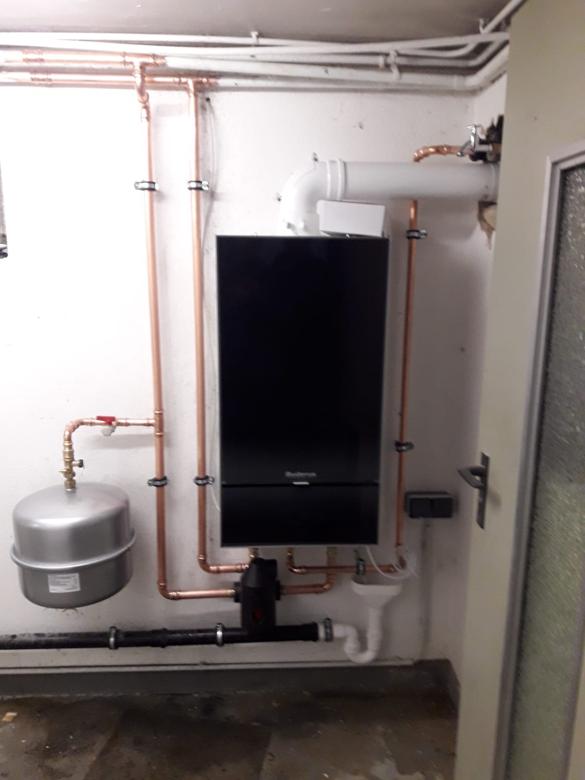 Zainstalowany nowoczesny piec centralnego ogrzewania marki Buderus z miedzianymi rurami, białym zbiornikiem wyrównawczym i odpływem kondensatu, zamocowany na białej ścianie w kotłowni.