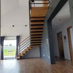 Nowoczesne, ażurowe schody z drewnianymi stopniami i czarną metalową balustradą w jasnym wnętrzu z widokiem na ogród przez okno.