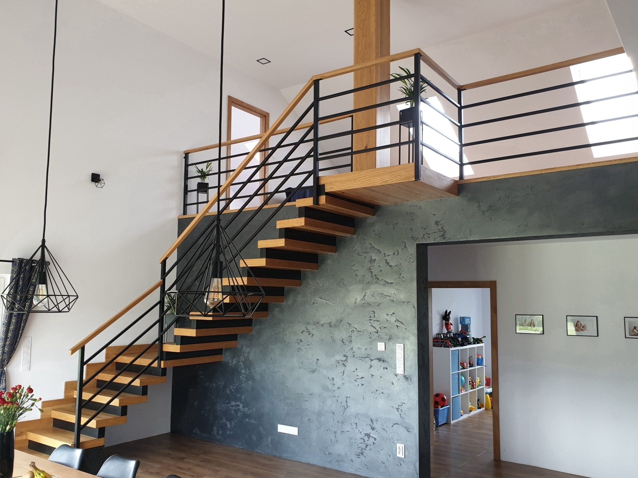 Nowoczesne schody drewniane z czarnymi metalowymi balustradami prowadzące na antresolę z widoczną rośliną w doniczce, oświetlone lampami wiszącymi w stylu industrialnym.