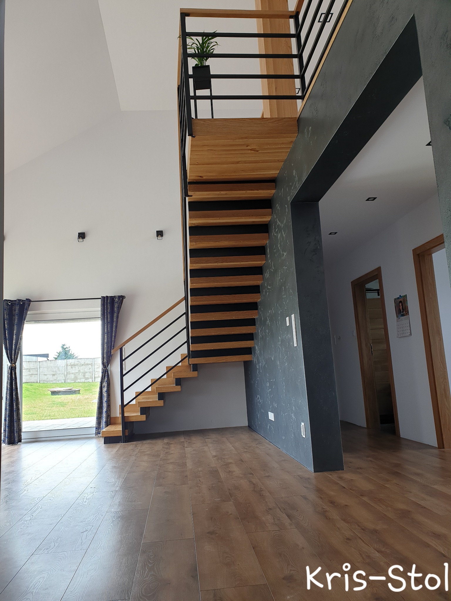 Nowoczesne, ażurowe schody z drewnianymi stopniami i czarną metalową balustradą w jasnym wnętrzu z widokiem na ogród przez okno.