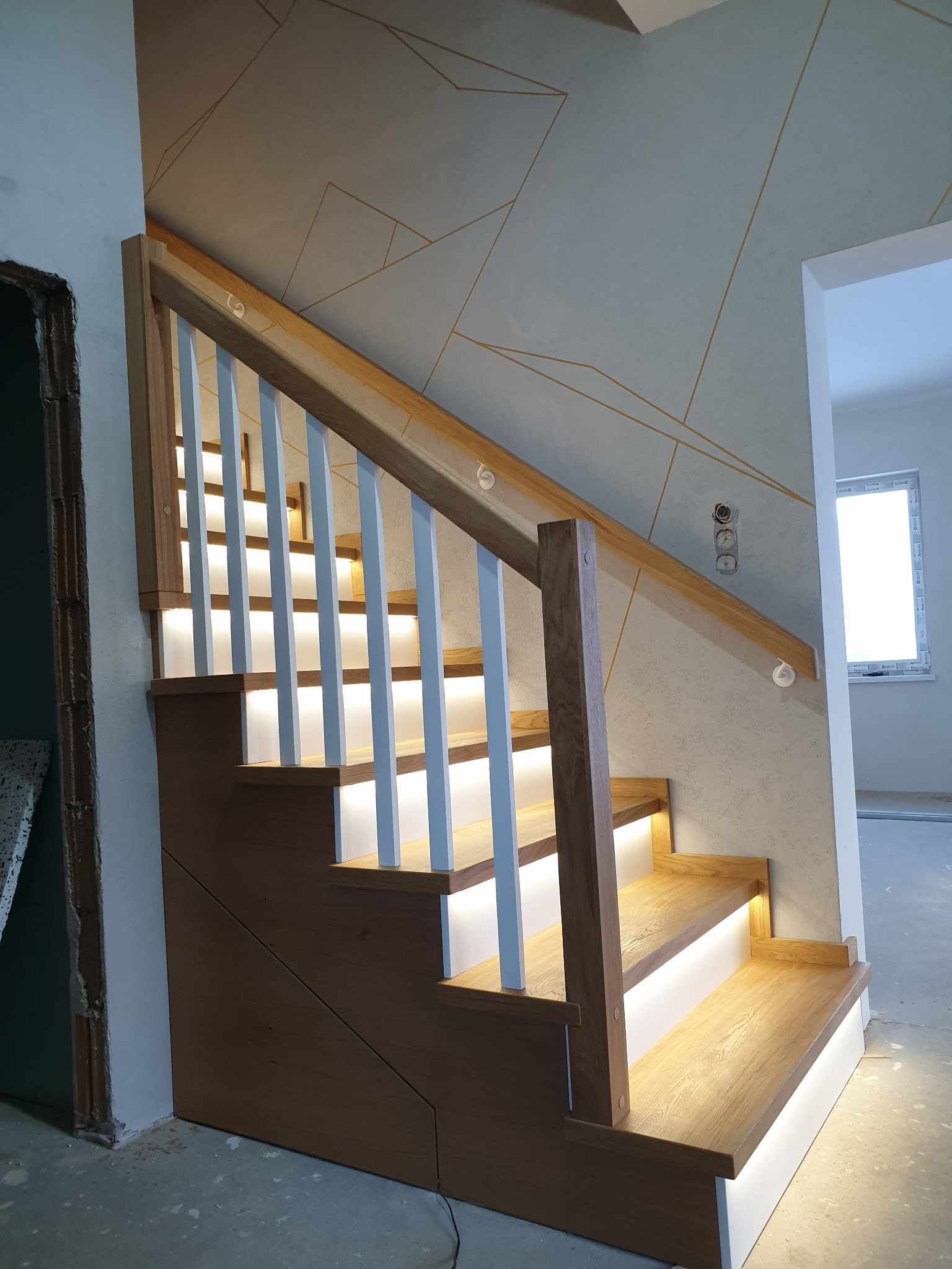 Nowoczesne schody z drewna dębowego z białymi tralkami i podświetleniem LED na każdym stopniu, widoczne gniazdka elektryczne na ścianie w tle.