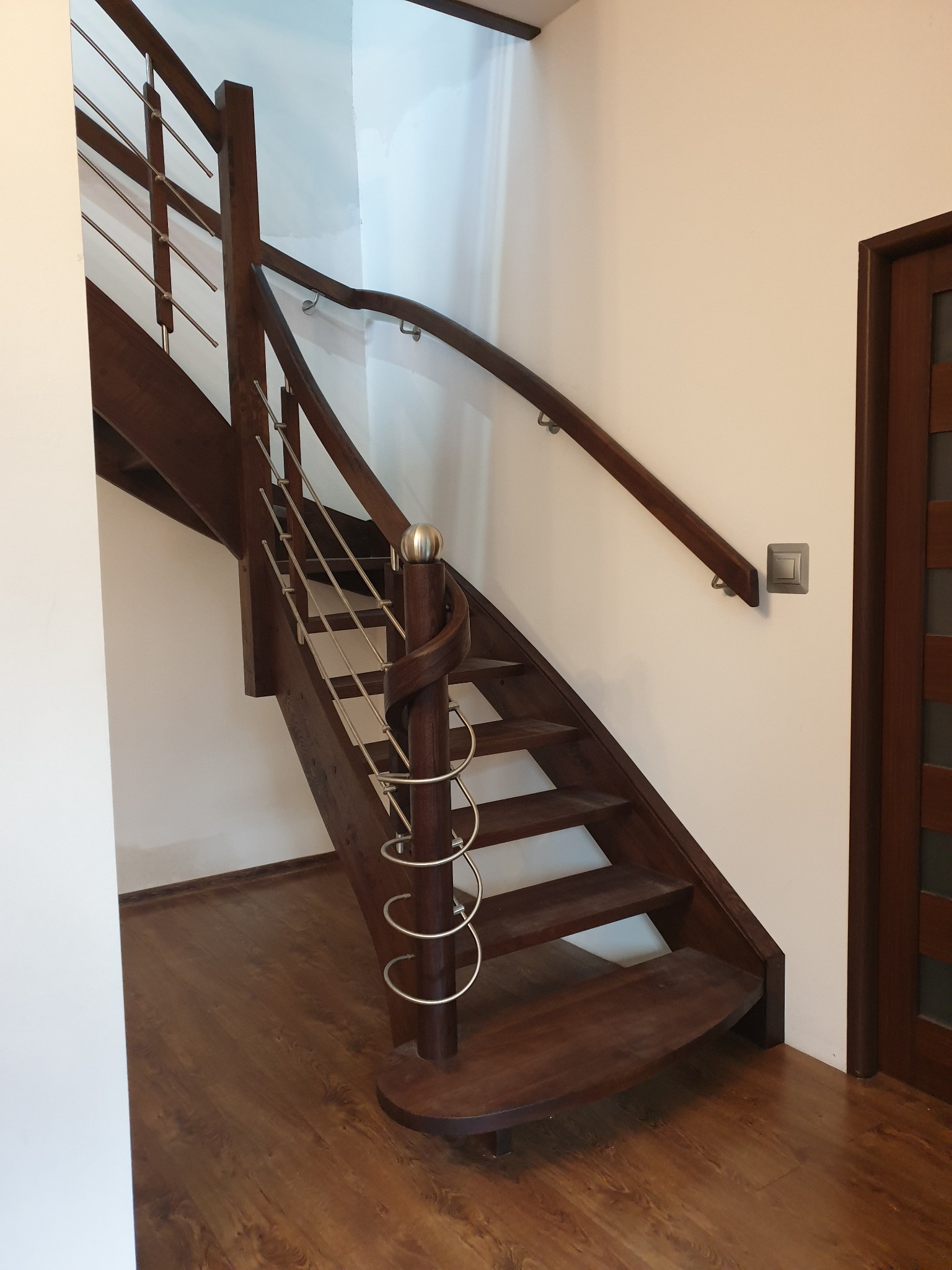 Ciemne, spiralne schody z metalowymi elementami balustrady i drewnianą poręczą, widoczne na tle białej ściany i drewnianej podłogi, z fragmentem drzwi po prawej stronie.