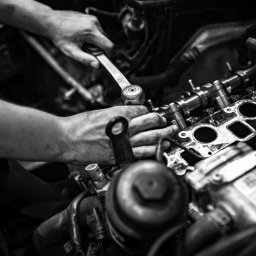 Automechanika Ratka DDCars - Czarno-białe zdjęcie przedstawia dłonie mechanika dokręcającego śruby na głowicy silnika za pomocą klucza dynamometrycznego. Widoczne elementy silnika oraz narzędzia w warsztacie. Ujęcie z bliska.
