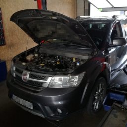 Automechanika Ratka DDCars - Czarny samochód marki Fiat na podnośniku w warsztacie z otwartą maską, widoczny silnik. Na podłodze widoczne narzędzia i pojemnik.