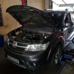 Czarny samochód marki Fiat na podnośniku w warsztacie z otwartą maską, widoczny silnik. Na podłodze widoczne narzędzia i pojemnik.