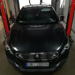 Automechanika Ratka DDCars - Czarny samochód osobowy marki Peugeot na podnośniku w warsztacie samochodowym, widok z przodu, tablica rejestracyjna zamazana.