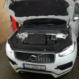 Automechanika Ratka DDCars - Białe Volvo XC90 z otwartą maską, widoczny silnik i akumulator w warsztacie.