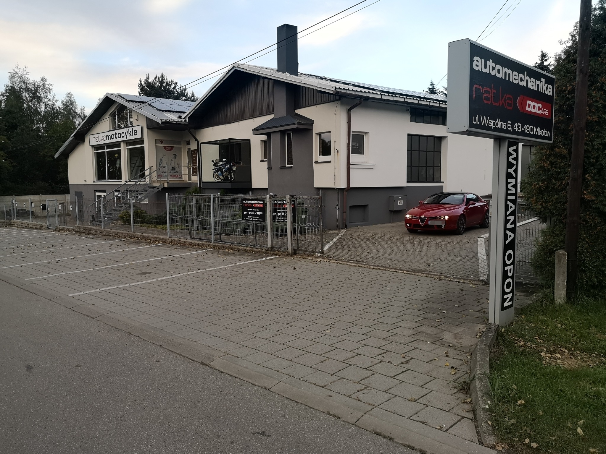 Budynek warsztatu samochodowego z szyldem 'Automechanika Ratka' i czerwonym samochodem Alfa Romeo zaparkowanym przed nim. Na budynku widoczne panele słoneczne, a w oknach wystawowych motocykle.