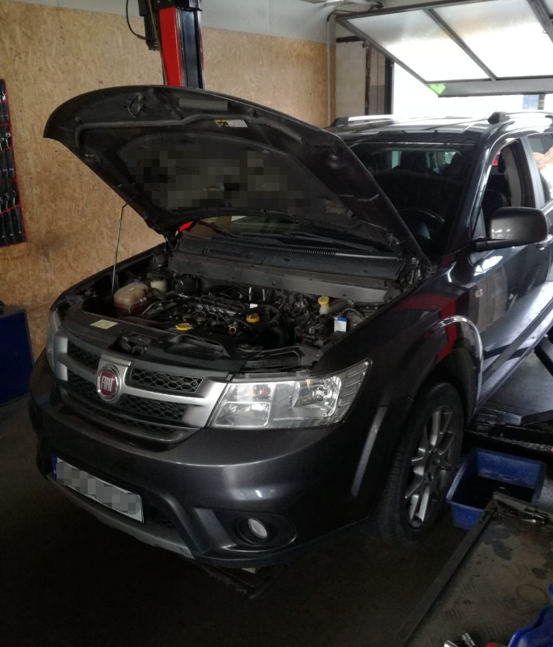 Czarny samochód marki Fiat na podnośniku w warsztacie z otwartą maską, widoczny silnik. Na podłodze widoczne narzędzia i pojemnik.