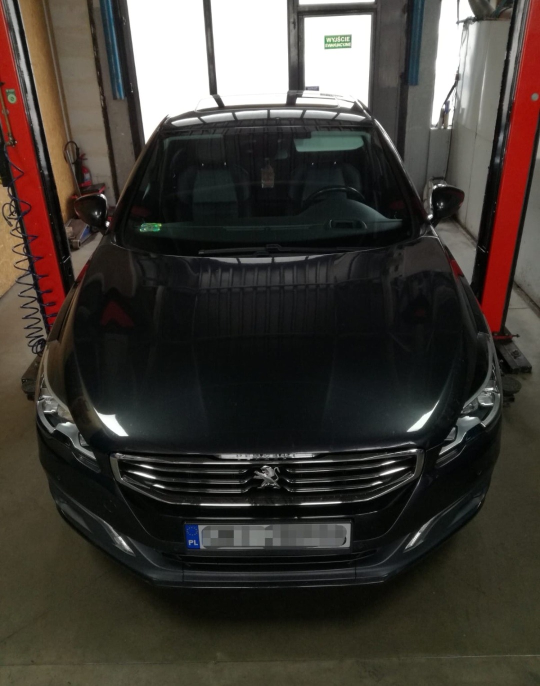 Czarny samochód osobowy marki Peugeot na podnośniku w warsztacie samochodowym, widok z przodu, tablica rejestracyjna zamazana.
