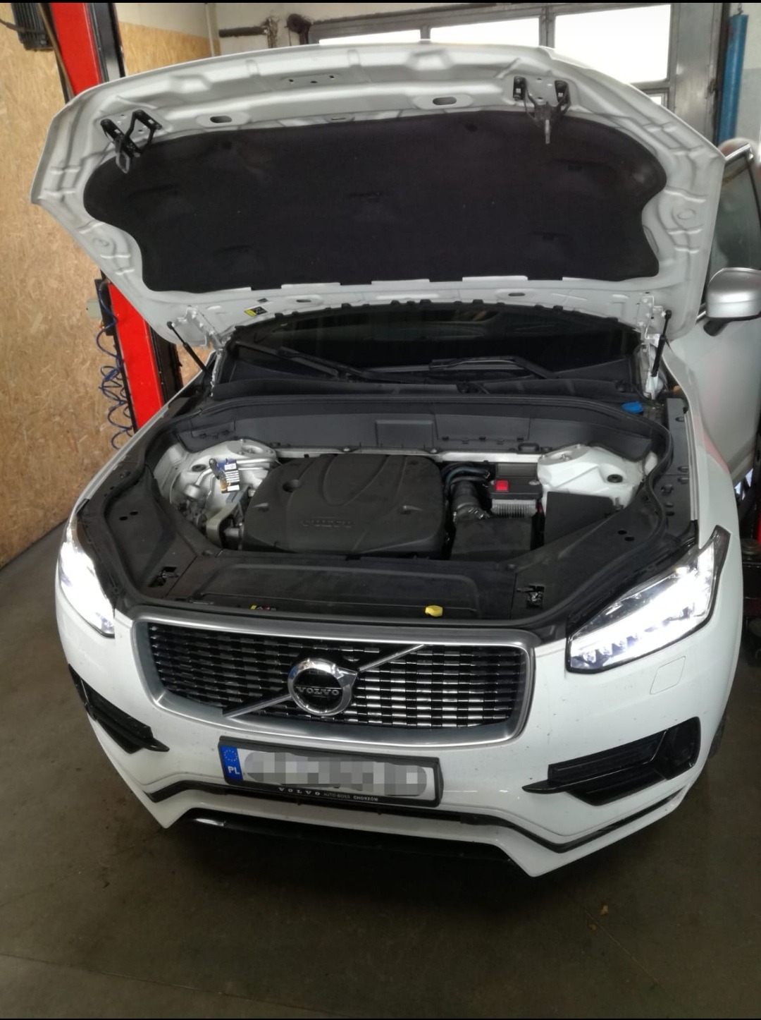 Białe Volvo XC90 z otwartą maską, widoczny silnik i akumulator w warsztacie.