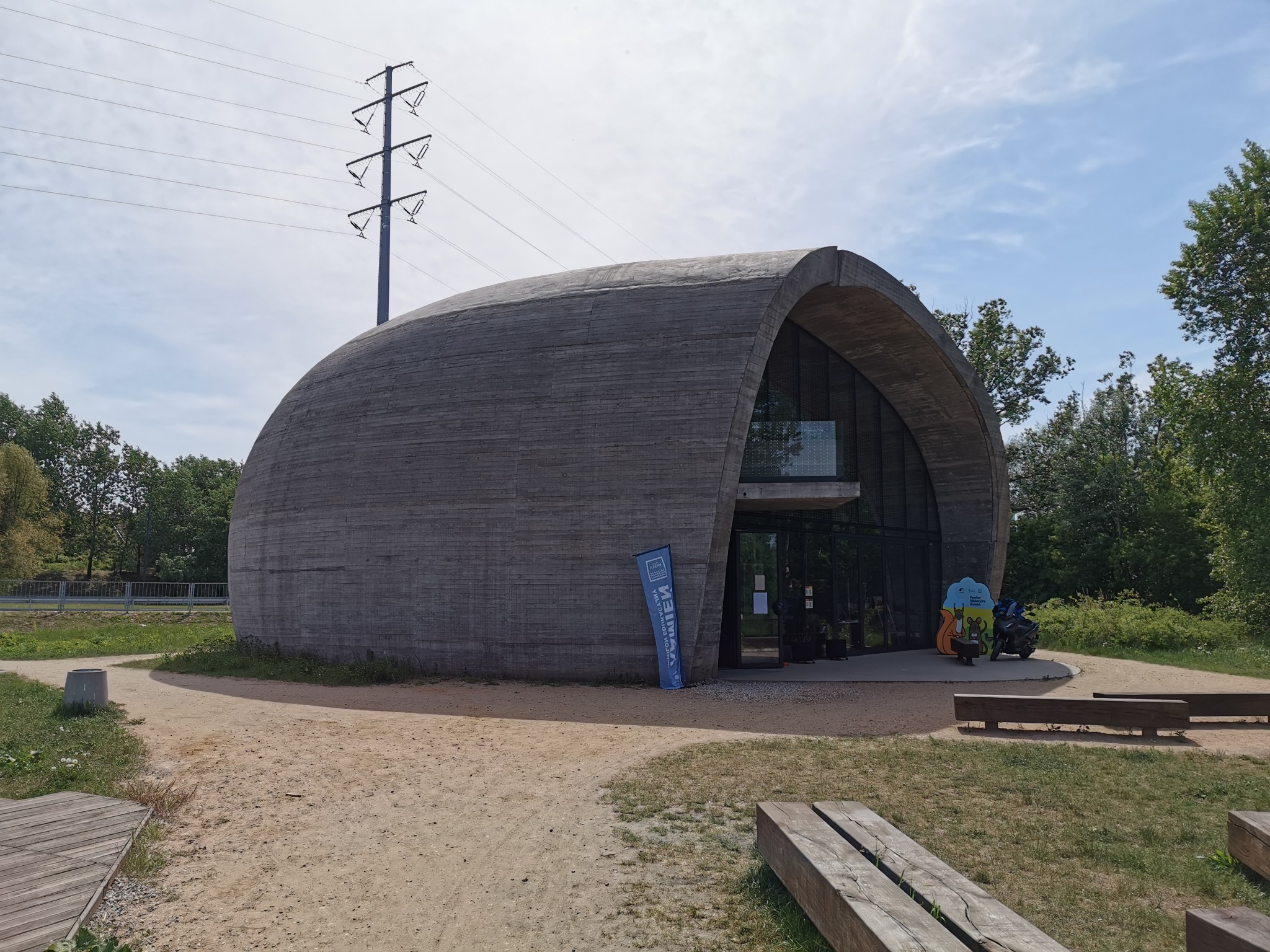 Pawilon Edukacyjny Kamień: betonowa konstrukcja w kształcie łuku z przeszklonym wejściem, otoczona piaszczystym terenem i zielenią, z widoczną linią wysokiego napięcia w tle.