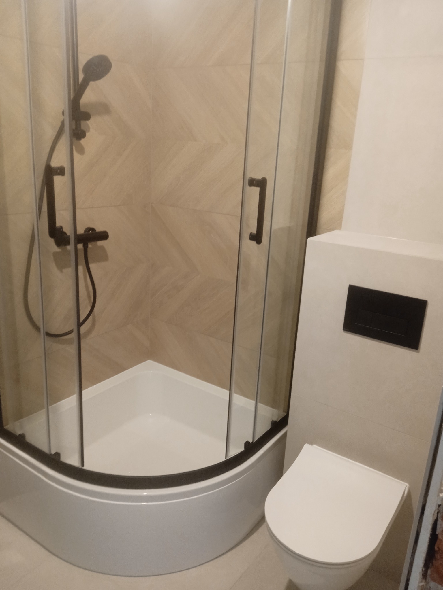 Wykończona łazienka: kabina prysznicowa z czarną armaturą i toaleta w zabudowie. Płytki w odcieniach beżu z delikatnym wzorem drewna. Nowoczesny design.