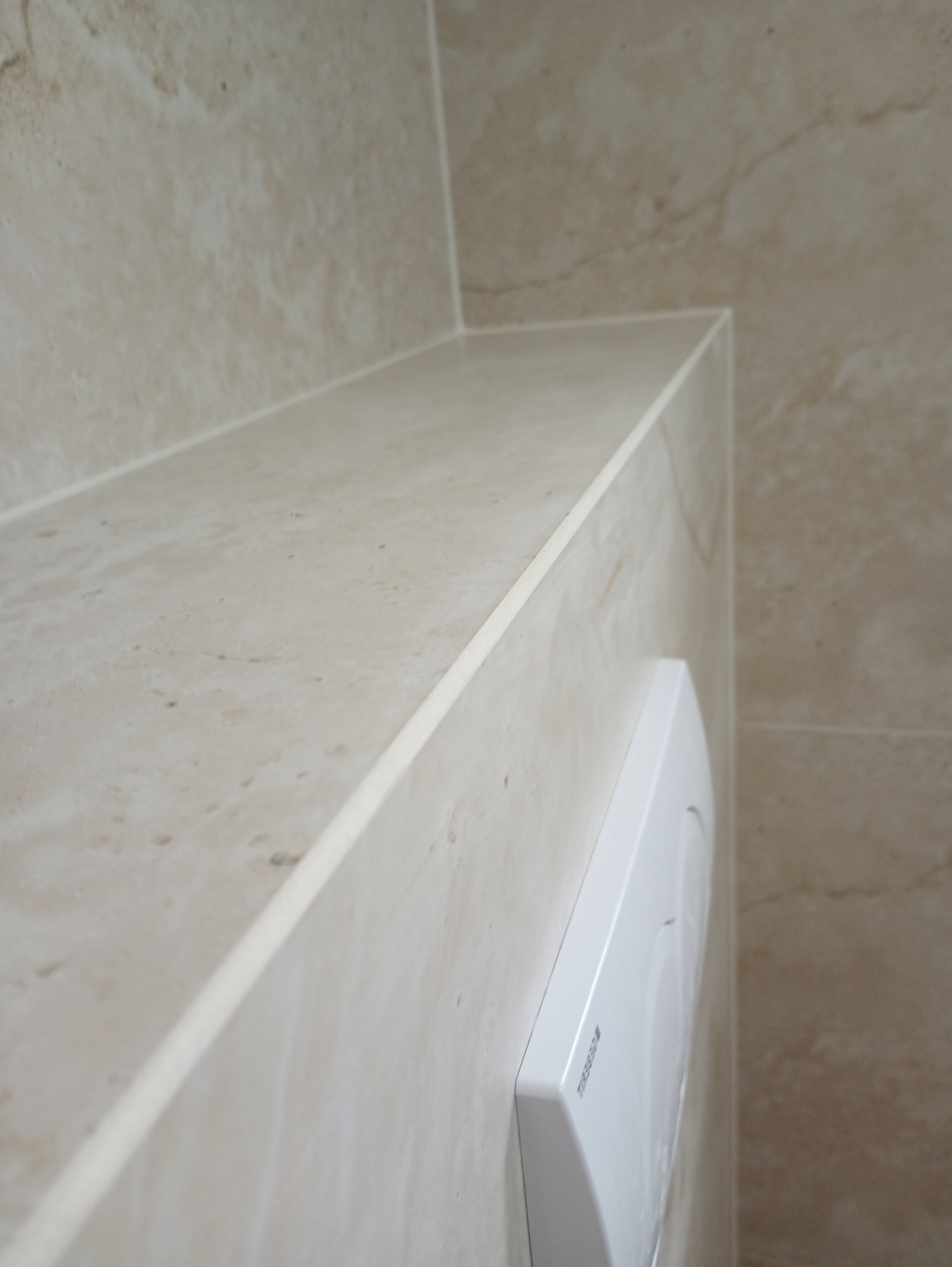 Beżowe płytki imitujące marmur, ułożone na ścianie i półce w łazience. Widoczny biały przycisk spłuczki. Precyzyjne fugi i kąty.