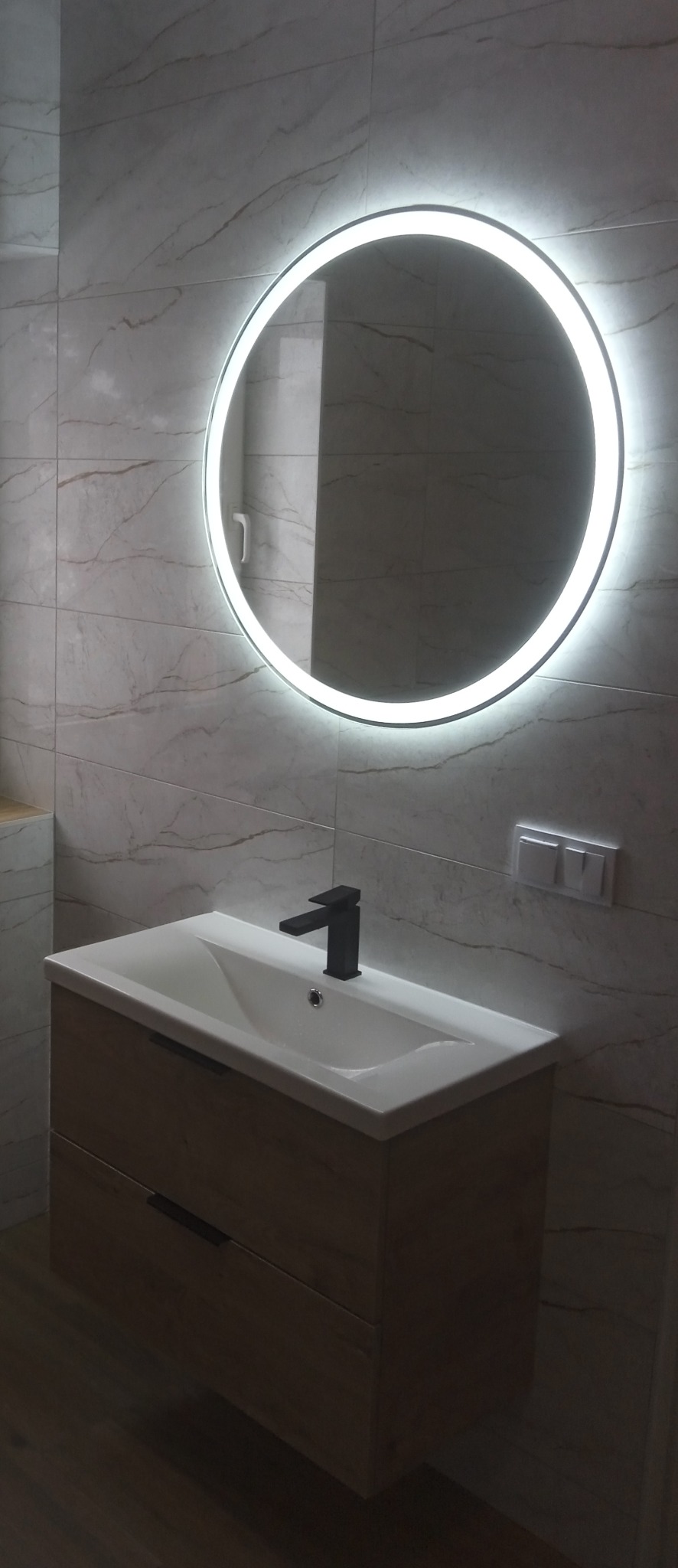 Nowoczesna łazienka: umywalka z szafką w odcieniach drewna, czarna bateria i okrągłe lustro z podświetleniem LED na tle marmurowych płytek.