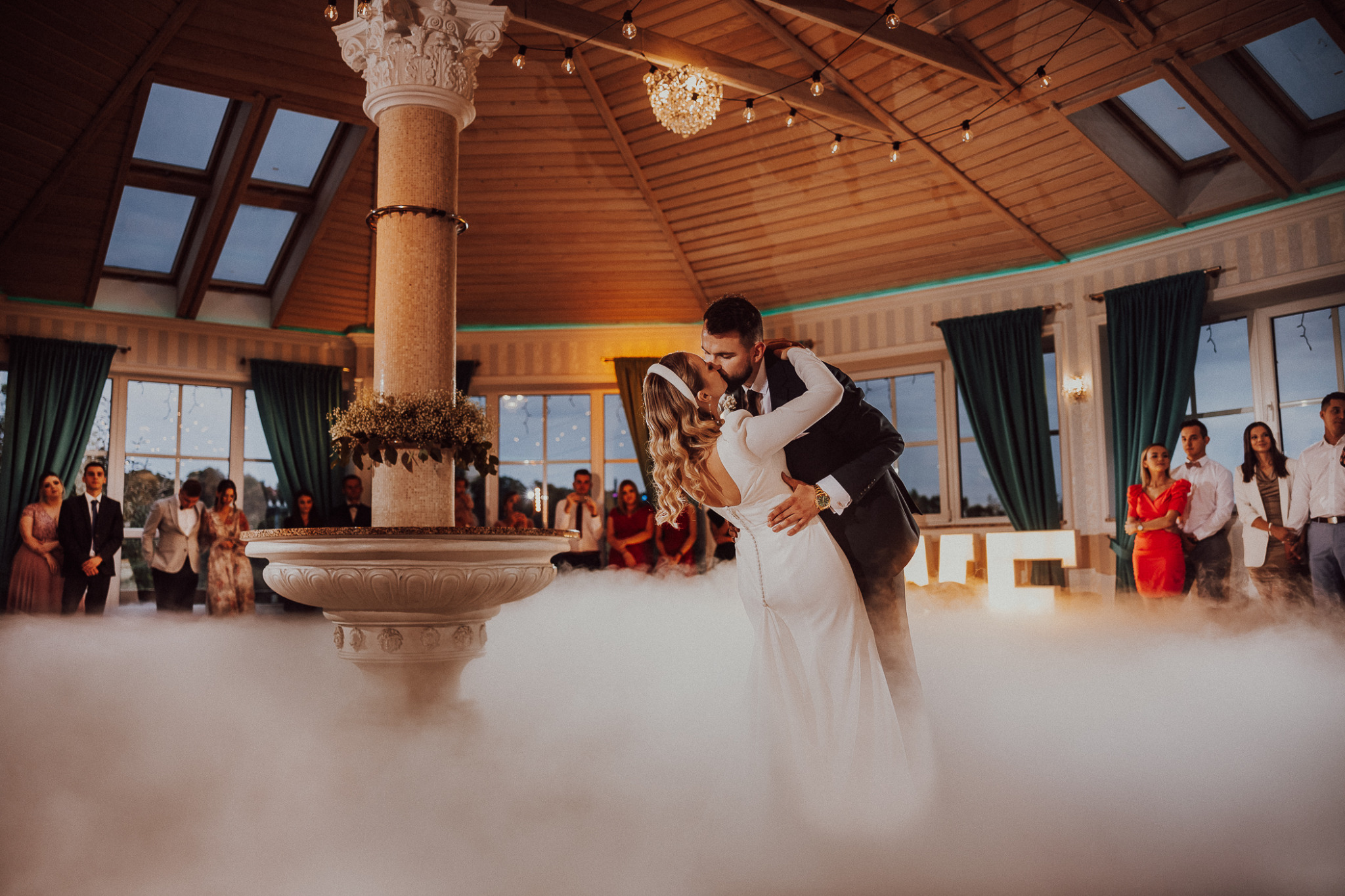 Pocałunek nowożeńców podczas pierwszego tańca w sali z kolumną, fontanną i ciężkim dymem, goście w tle.