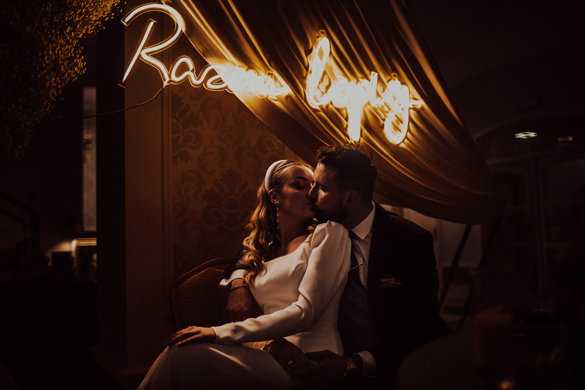 Pocałunek pary młodej siedzącej na krześle, podświetlony neonowym napisem i drapowanym materiałem w ciepłych odcieniach.