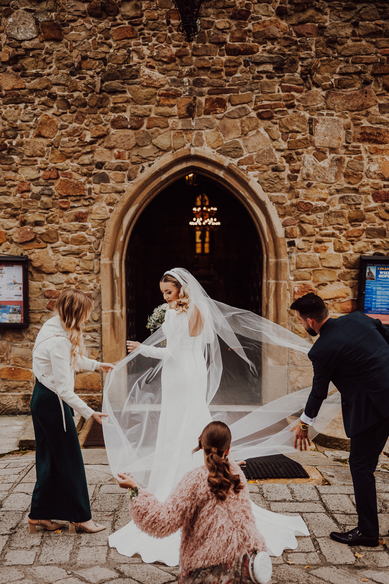 Panna młoda w białej sukni i welonie wychodzi z kamiennego kościoła, asystują jej dwie kobiety i mężczyzna, poprawiając welon.