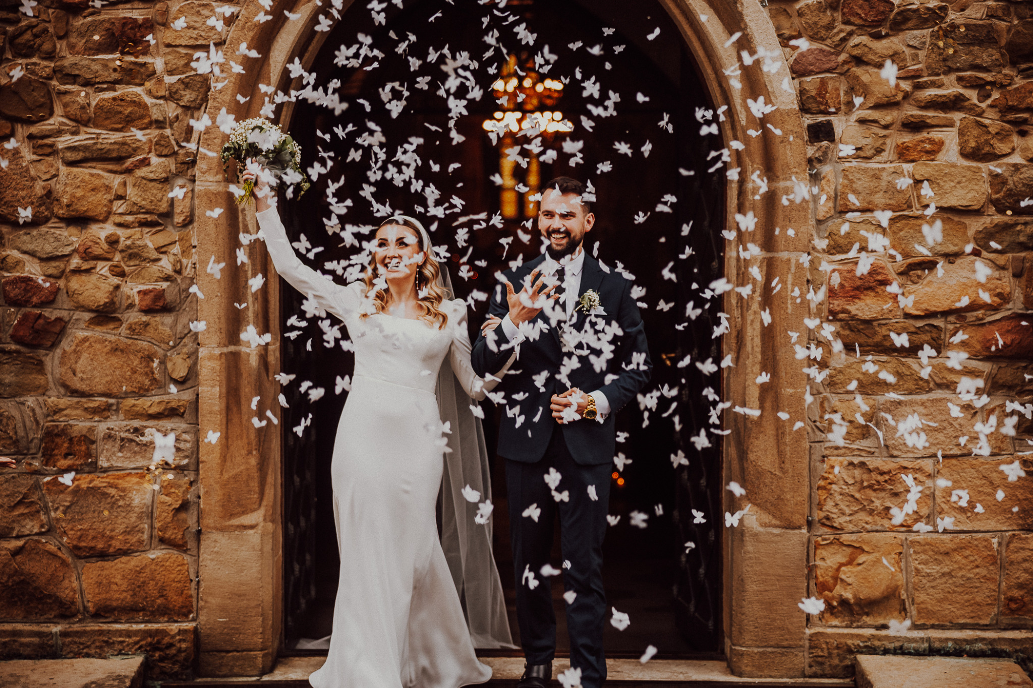 Uśmiechnięta para młoda wychodzi z kamiennego kościoła, obsypywana białymi konfetti w kształcie motyli, panna młoda trzyma bukiet w uniesionej dłoni.