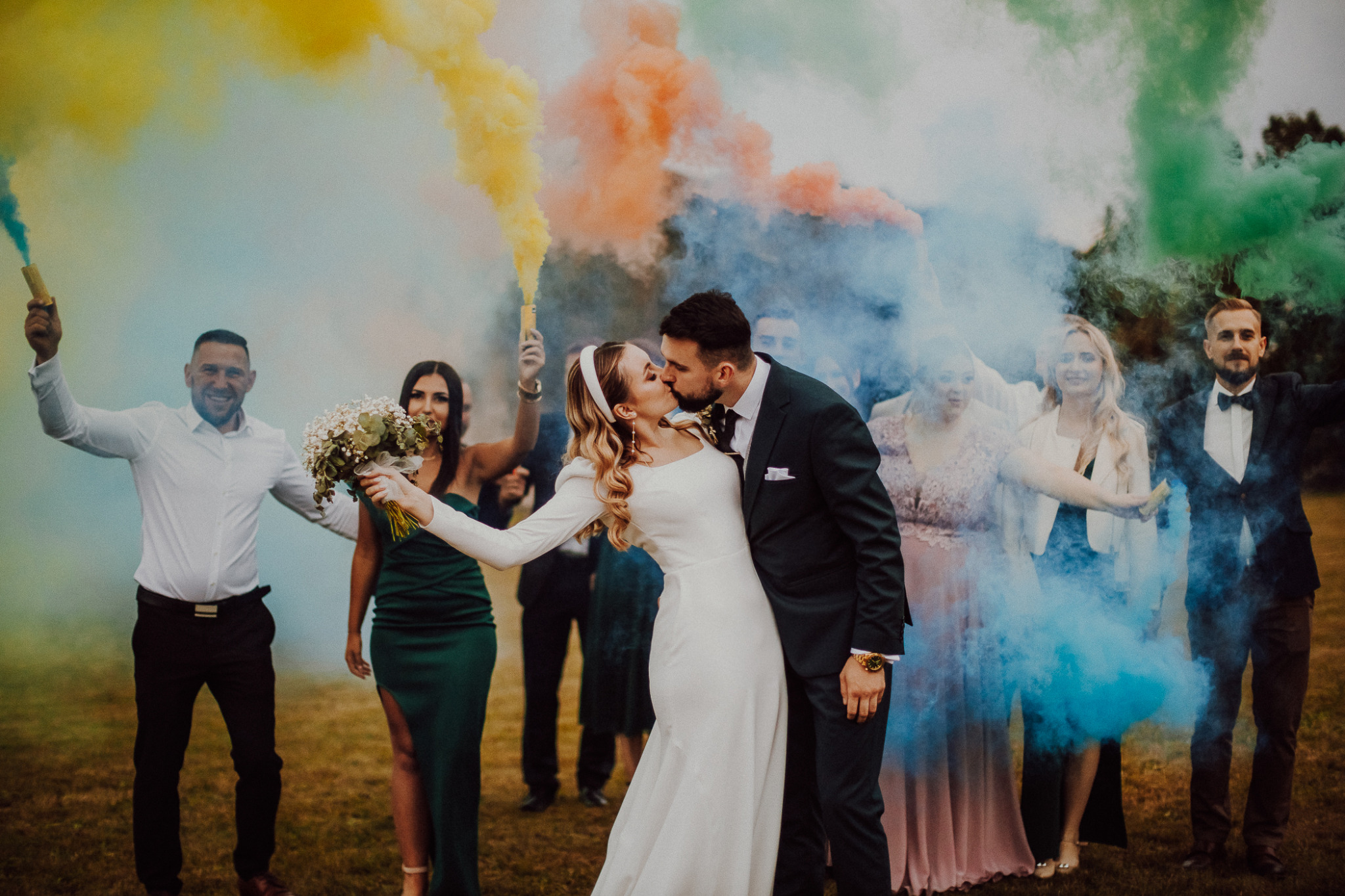 Pocałunek nowożeńców na tle kolorowych dymów trzymanych przez gości weselnych na łące.