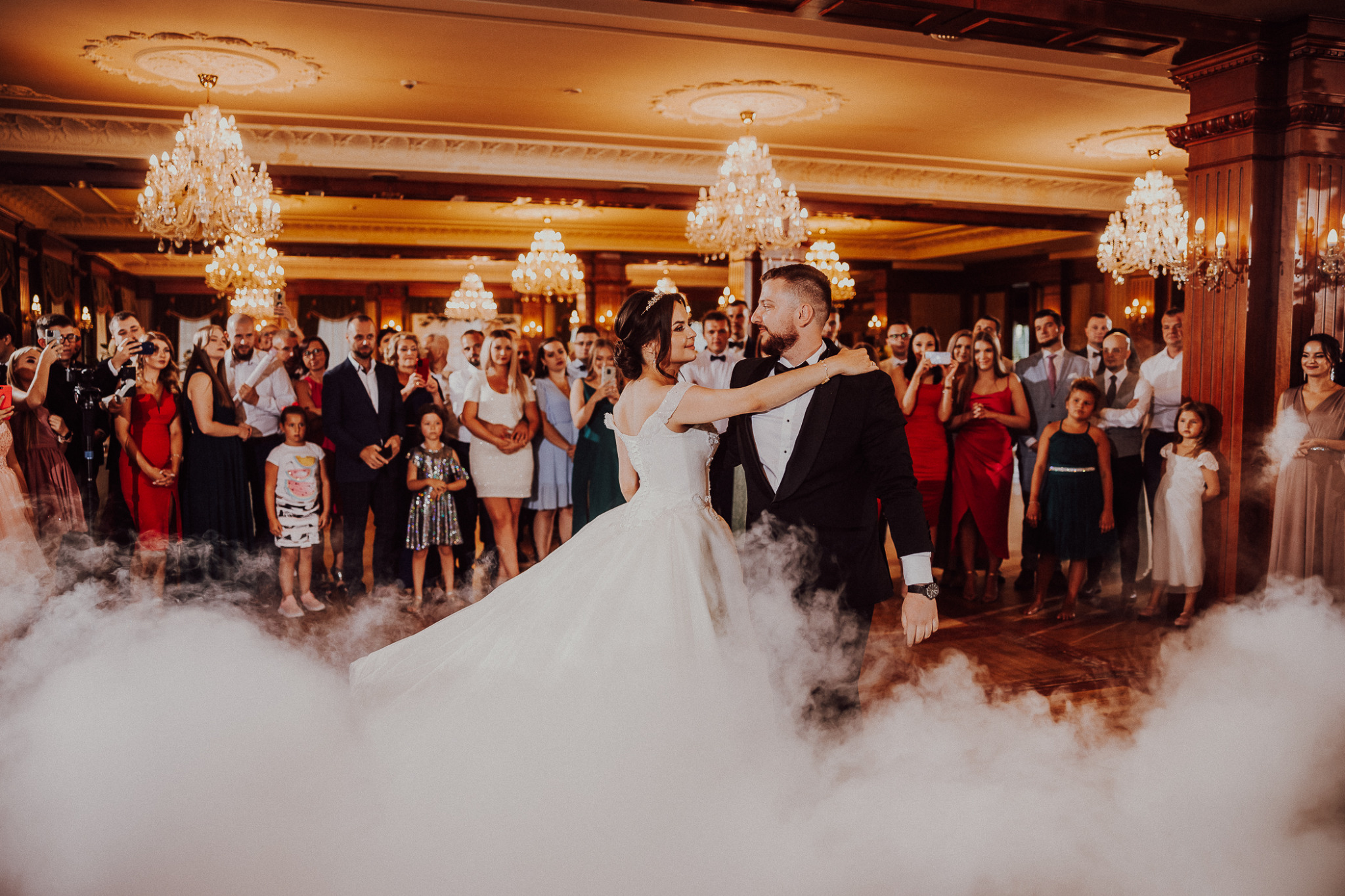 Para młoda w tańcu podczas wesela, otoczona sztucznym dymem, z gośćmi obserwującymi w tle w bogato zdobionej sali balowej z kryształowymi żyrandolami.
