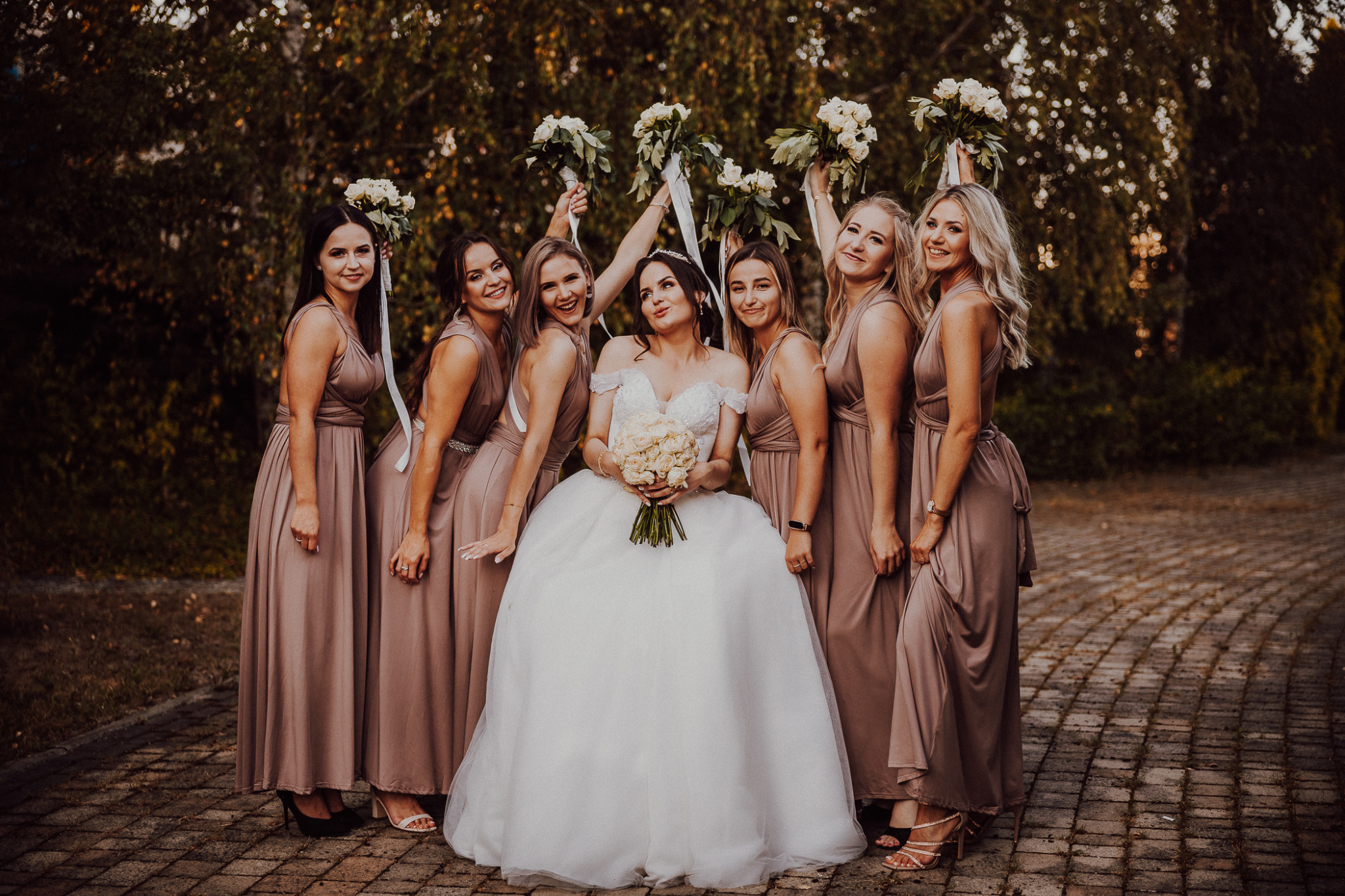 Panna młoda w białej sukni z bukietem róż otoczona druhnami w długich, brązowych sukienkach, trzymającymi bukiety nad głowami, pozująca na brukowanej ścieżce w plenerze.
