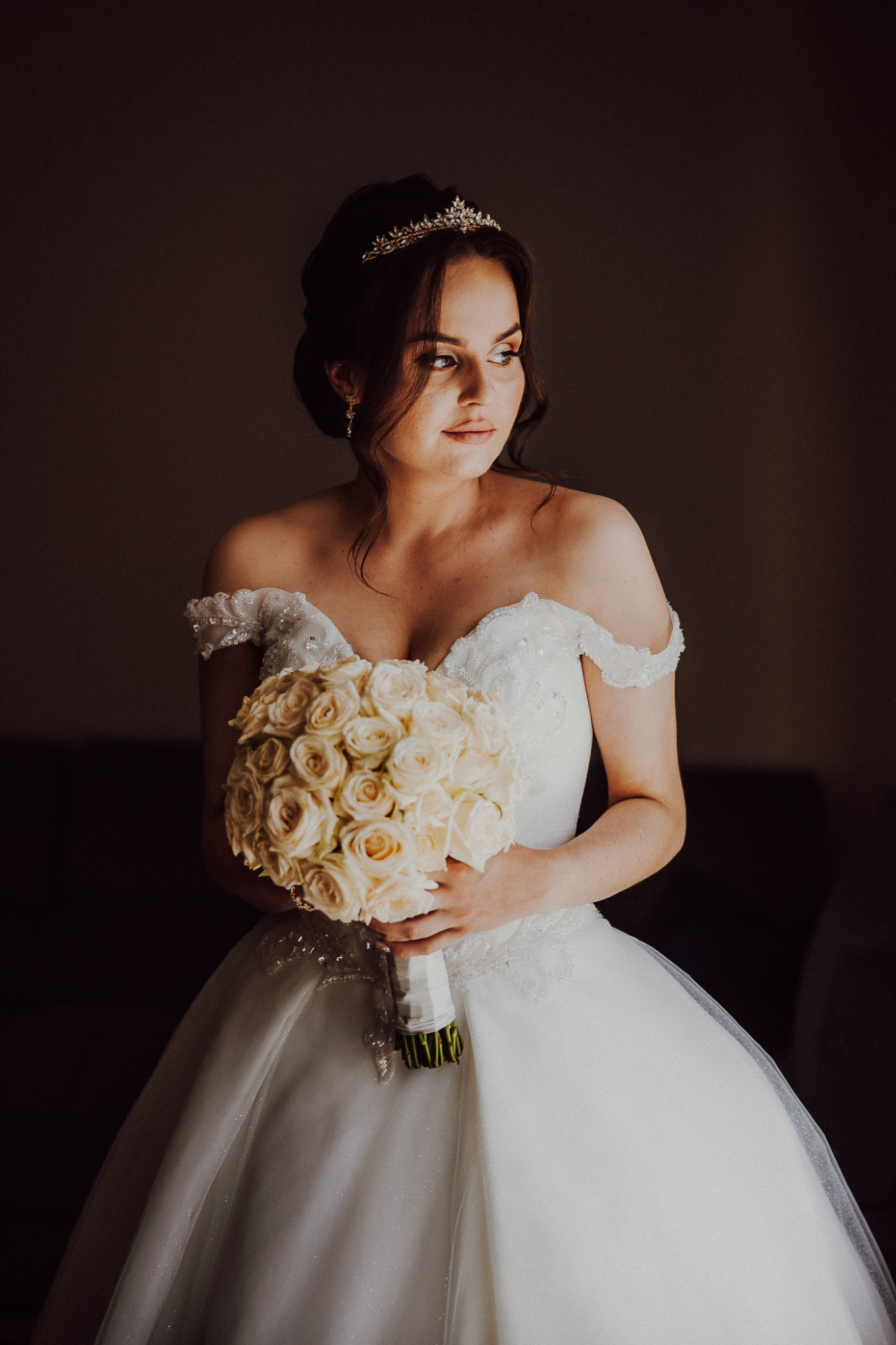 Panna młoda w białej sukni z odkrytymi ramionami, trzymająca bukiet kremowych róż i spoglądająca w bok, z tiarą na włosach.