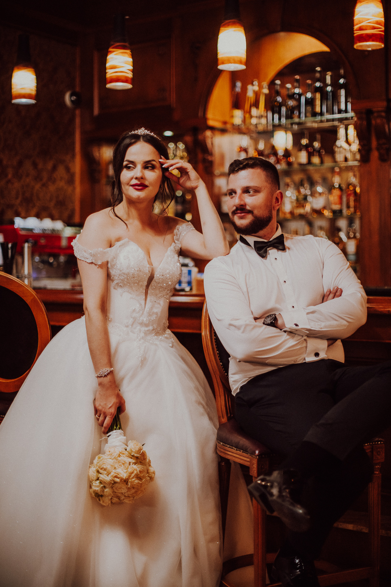 Panna młoda w bogato zdobionej sukni i tiarze, trzymająca bukiet kremowych róż, pozuje obok pana młodego w muszce, siedzących na barowych krzesłach na tle baru z oświetlonymi półkami pełnymi butelek.