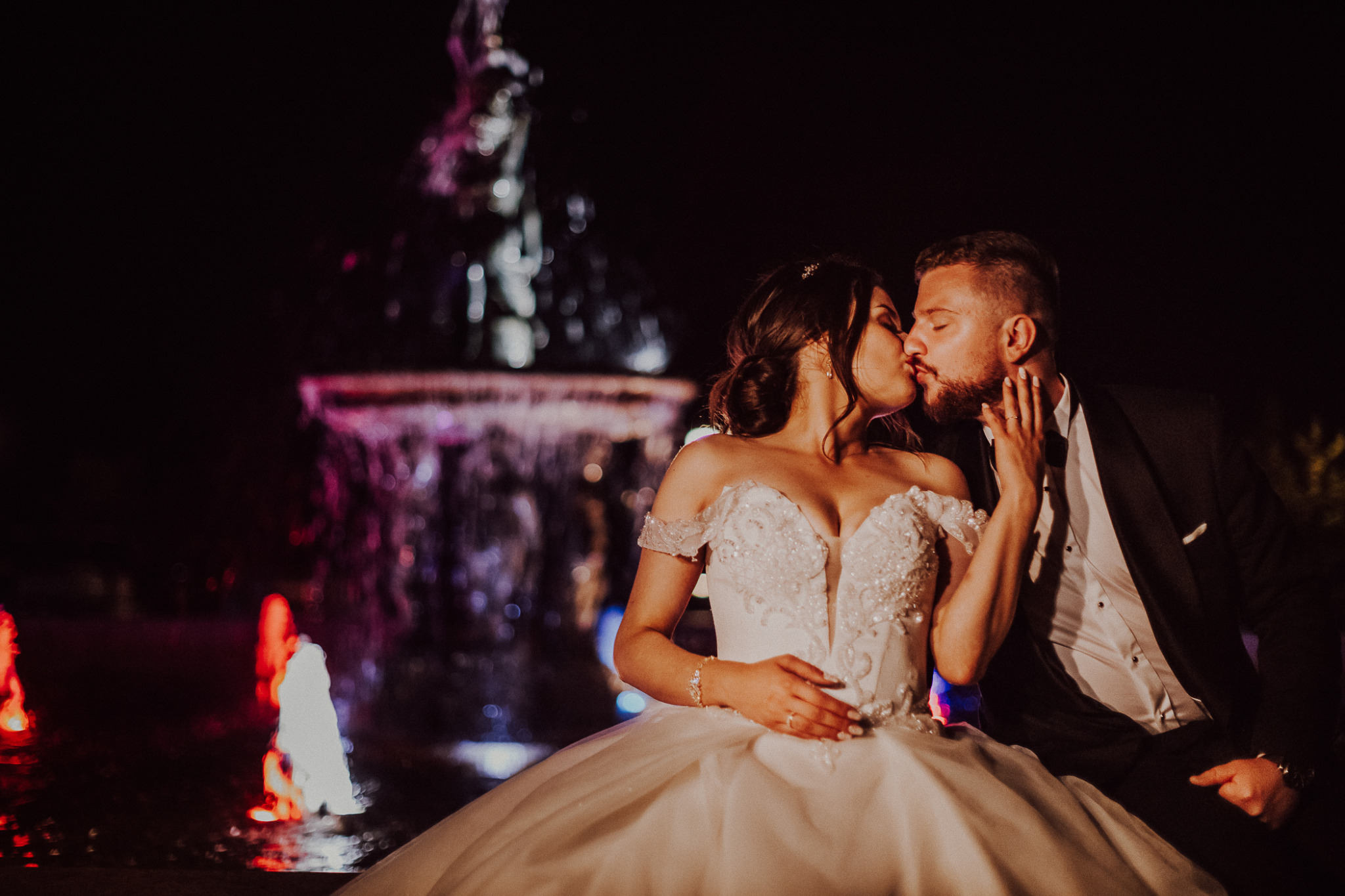 Pocałunek nowożeńców na tle podświetlanej fontanny nocą, panna młoda w sukni z odkrytymi ramionami i zdobieniami, pan młody w czarnym smokingu.