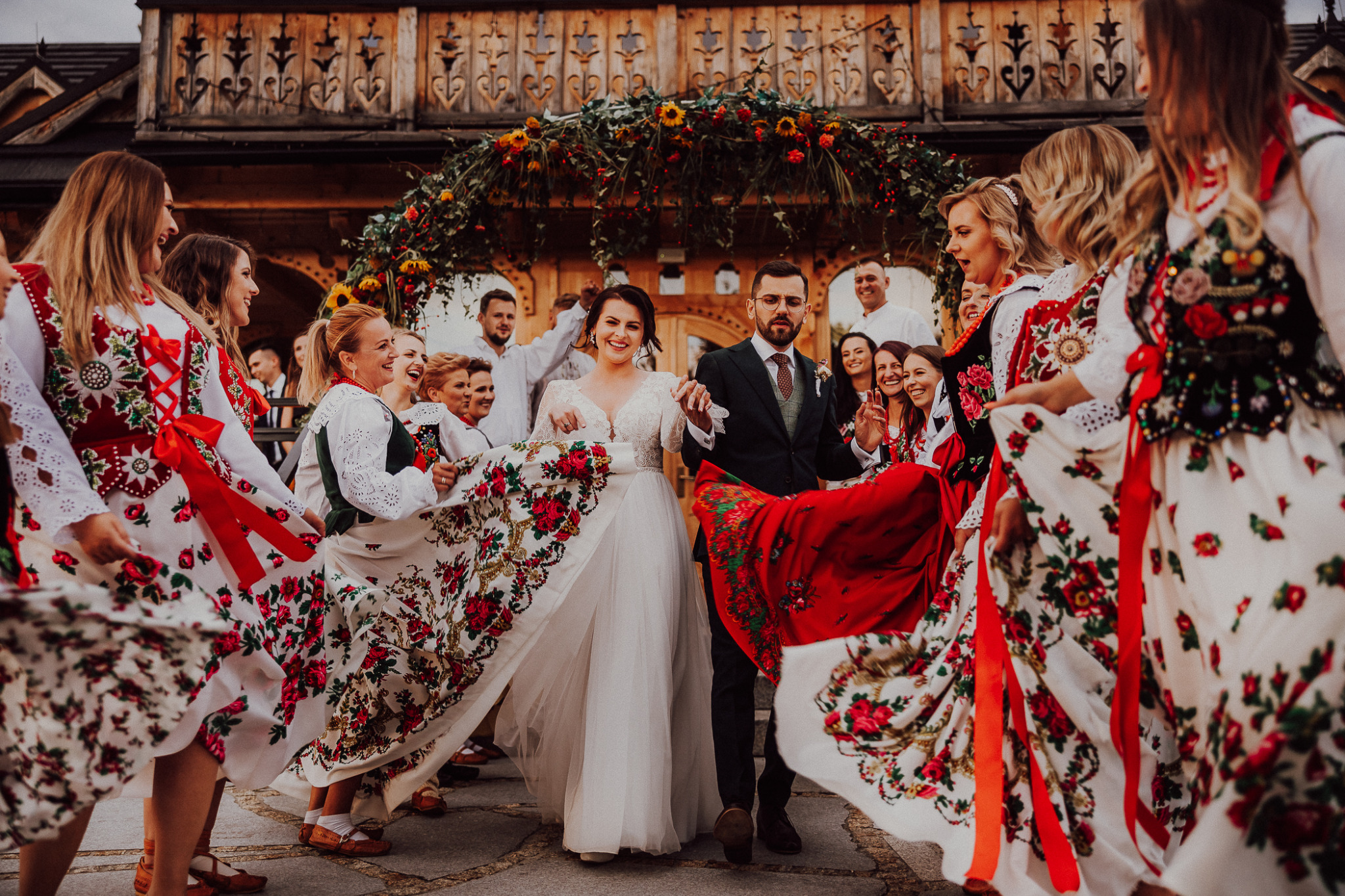 Para młoda w strojach ślubnych prowadzona przez gości weselnych w tradycyjnych, ludowych strojach przed drewnianym budynkiem z dekoracją kwiatową.