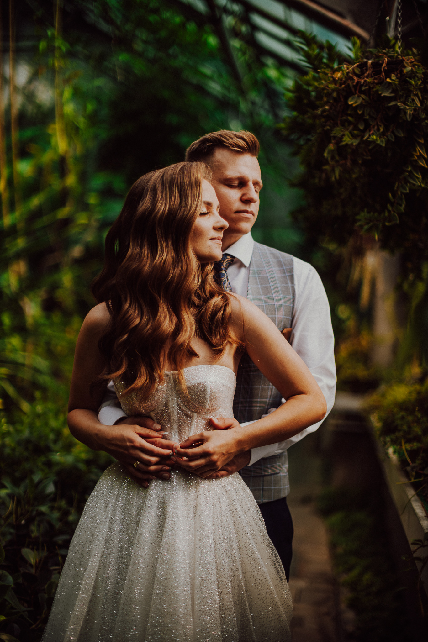 Para młoda w objęciach w szklarni, panna młoda w brokatowej sukni bez ramiączek, pan młody w kraciastej kamizelce, oboje z zamkniętymi oczami, otoczeni zielenią.