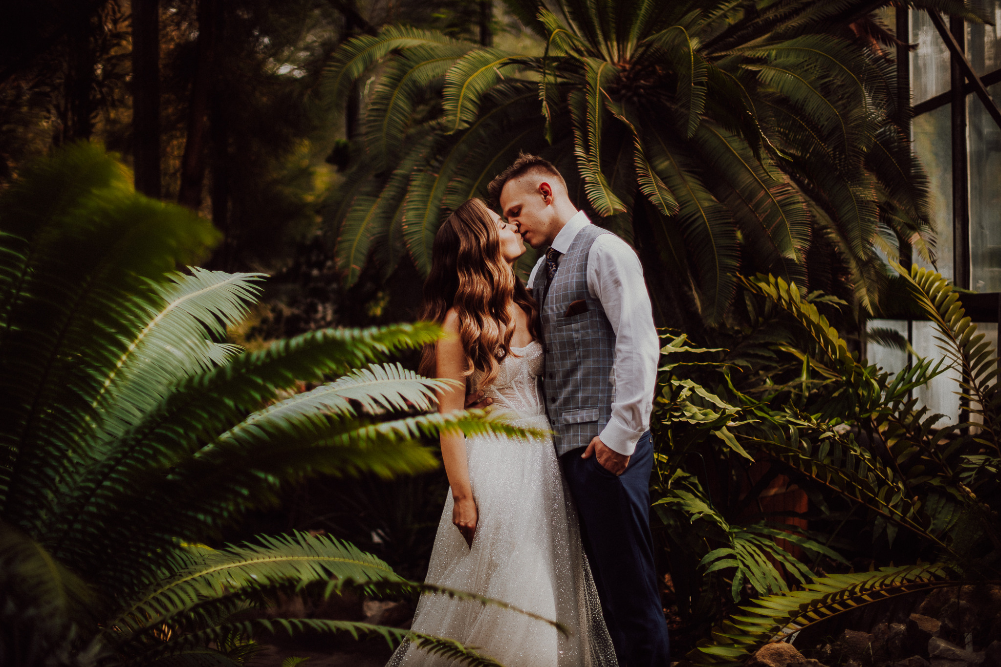 Para młoda w objęciach, pocałunek na tle bujnej zieleni w szklarni, panna młoda w błyszczącej sukni, pan młody w kraciastej kamizelce.