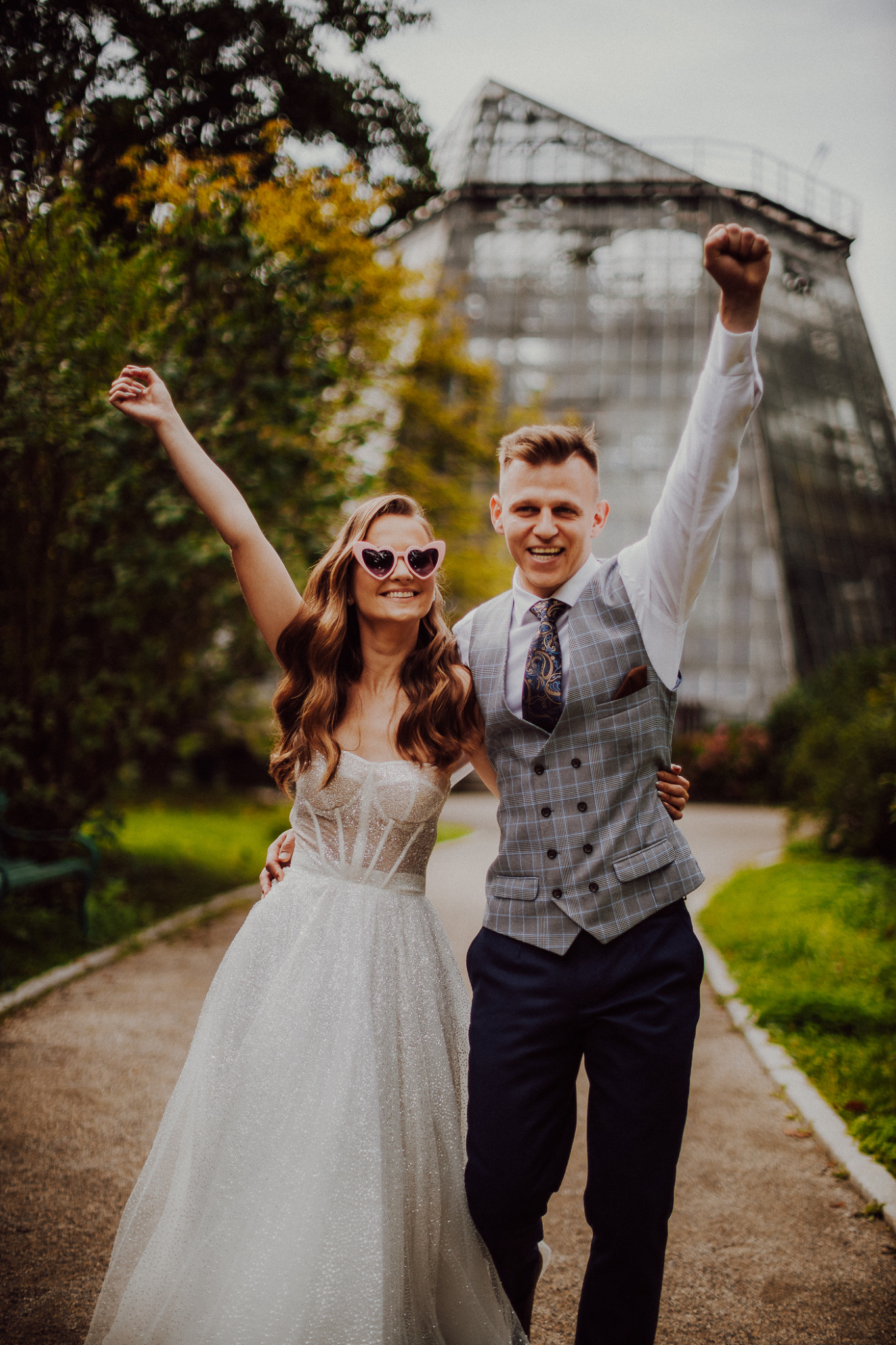 Uśmiechnięta para młoda świętuje na ścieżce w parku, trzymając ręce w górze; panna młoda w okularach w kształcie serc, w tle szklarnia.