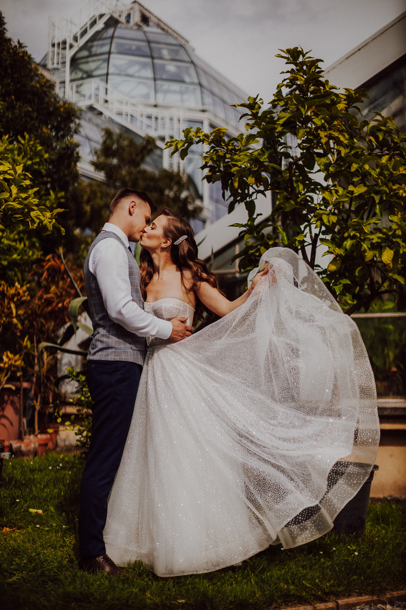 Pocałunek pary młodej na tle szklarni, panna młoda w sukni z brokatem, delikatnie unosi welon.