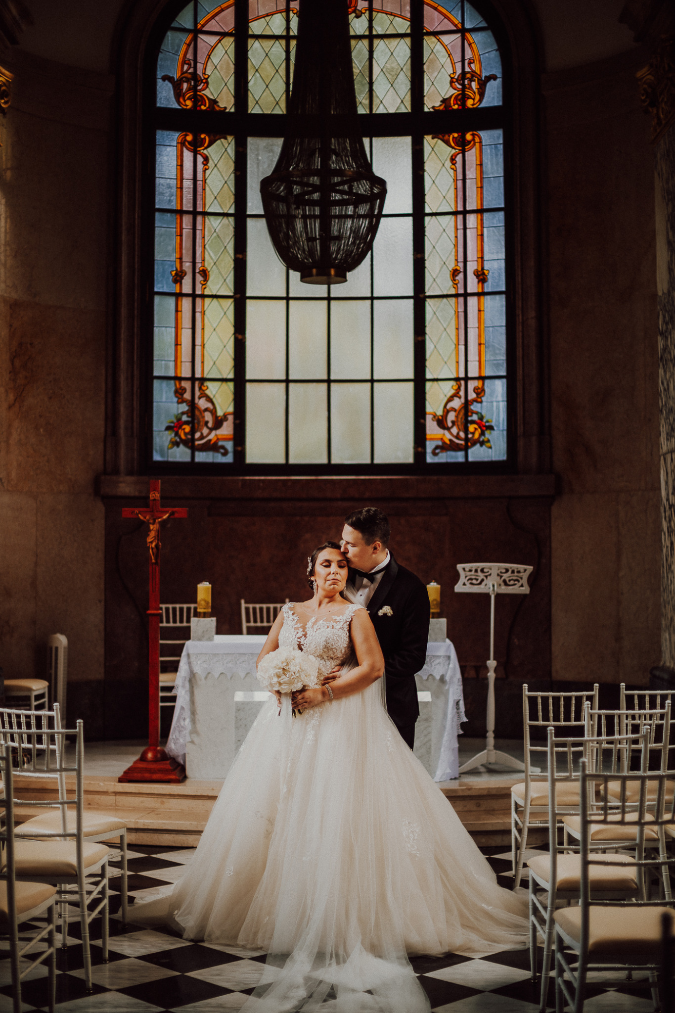 Para młoda w kościele: pan młody w czarnym smokingu całuje pannę młodą w sukni balowej z koronkową górą, trzymającą bukiet białych kwiatów, na tle witrażu i żyrandola.