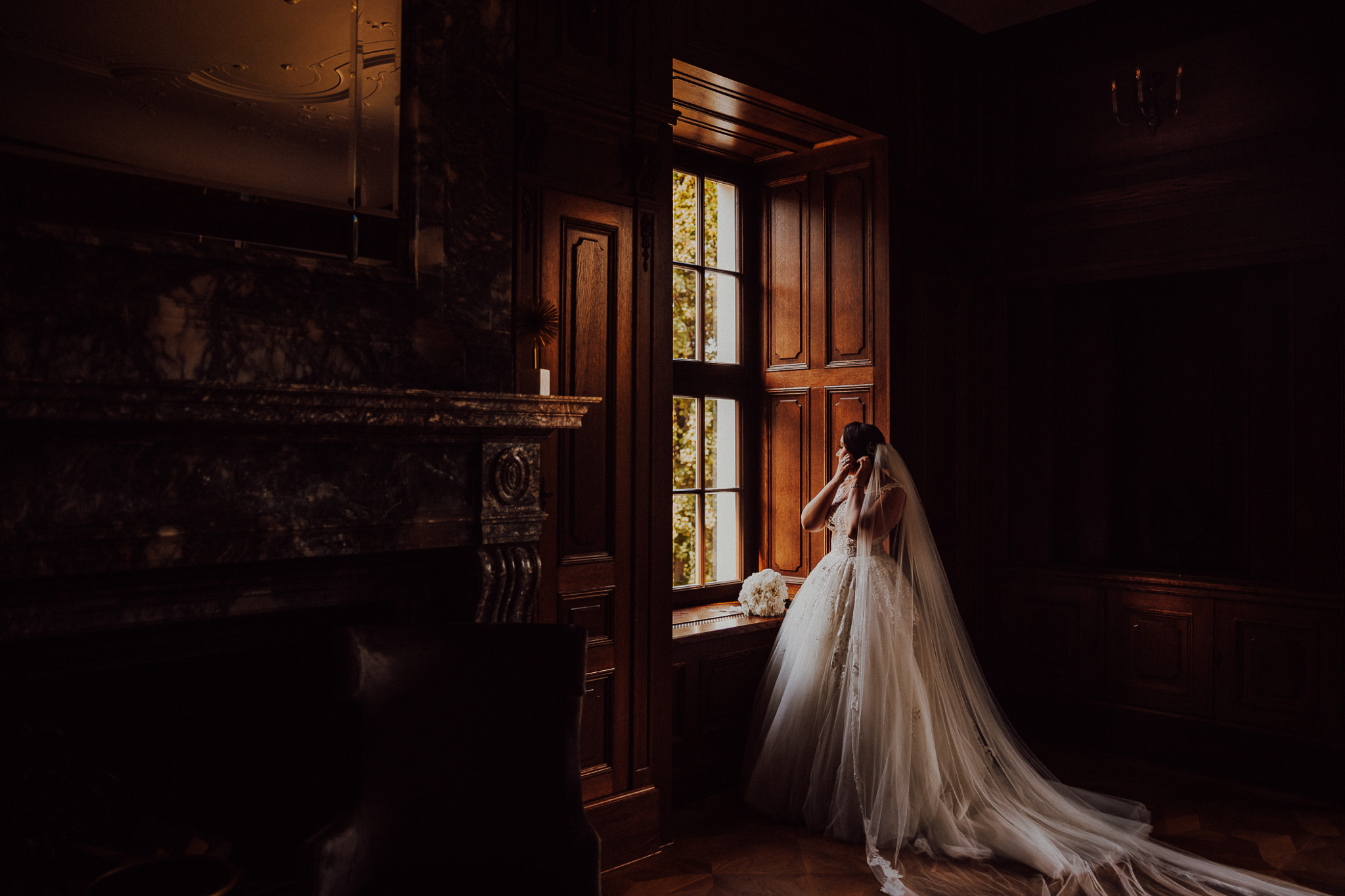 Panna młoda w koronkowej sukni i welonie, stojąca przy drewnianym oknie w ciemnym wnętrzu z bukietem białych kwiatów, poprawiająca kolczyk