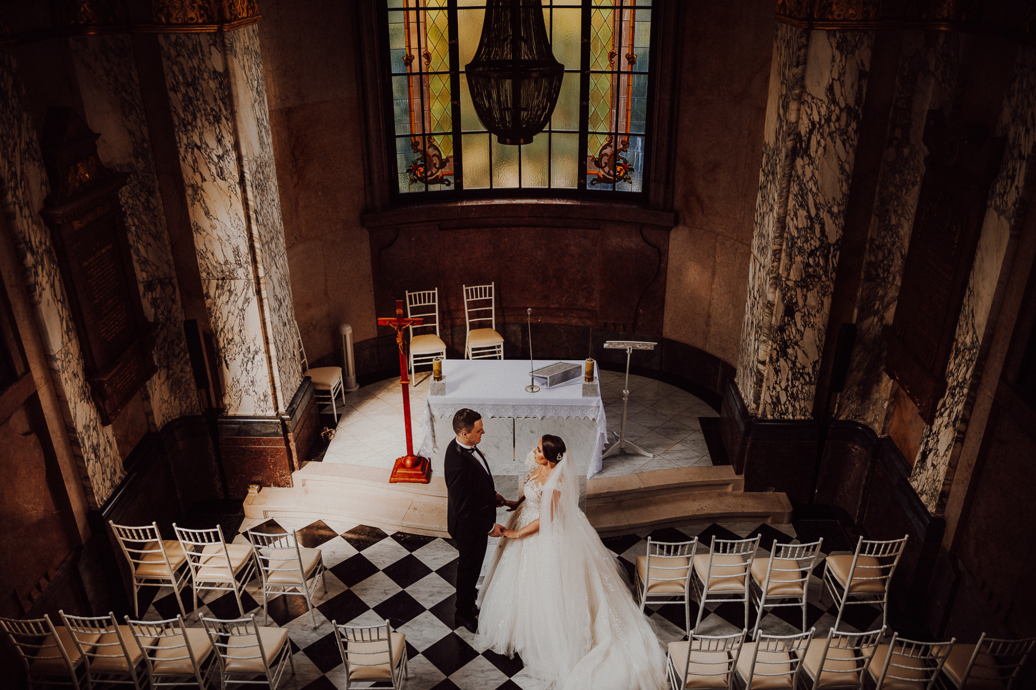 Para młoda trzymająca się za ręce przed ołtarzem w kościele z marmurowymi kolumnami i witrażem w tle, widok z góry.
