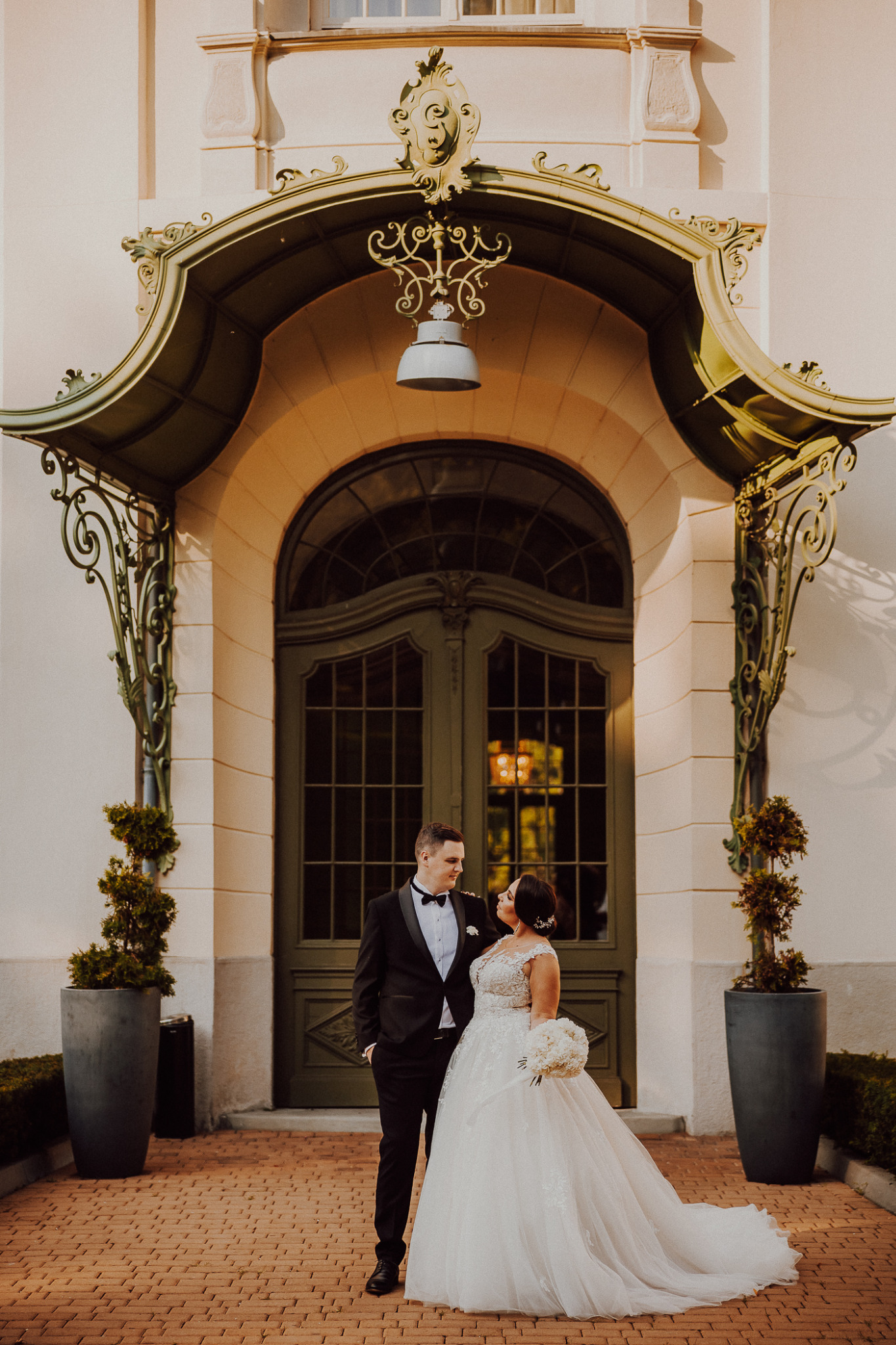 Para młoda w eleganckich strojach stoi przed ozdobnym wejściem z zielonymi drzwiami i kutą konstrukcją, panna młoda trzyma bukiet białych kwiatów.