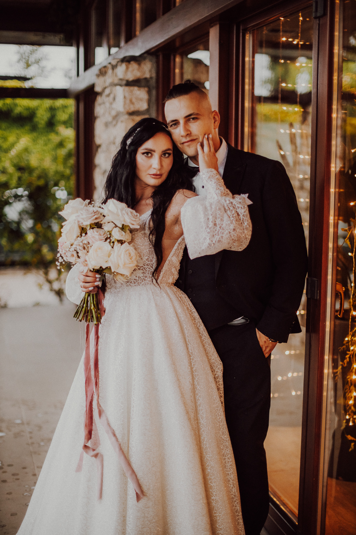 Elegancka para młoda w strojach ślubnych pozuje przed przeszklonym wejściem z dekoracyjnymi światełkami. Panna młoda trzyma bukiet różowych i białych kwiatów z zwisającymi wstążkami.