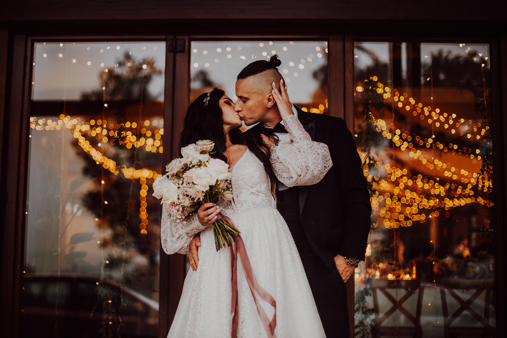 Pocałunek nowożeńców na tle okna udekorowanego światełkami, panna młoda w koronkowej sukni z bukietem białych róż, pan młody w czarnym smokingu z oryginalną fryzurą.