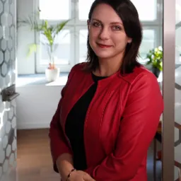 Portret kobiety w czerwonej skórzanej kurtce na tle jasnego biura z roślinami doniczkowymi i geometrycznym wzorem na szklanych drzwiach.