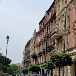 Ulica z odnowionymi kamienicami w różnych odcieniach cegły i beżu, z małymi balkonami z kutą balustradą, oświetlona przez latarnię uliczną w stylu retro.