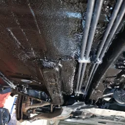 Podwozie samochodu po zabezpieczeniu antykorozyjnym, widoczne elementy zawieszenia i przewody hamulcowe, zabezpieczone czarną, błyszczącą powłoką, uniesione na podnośniku warsztatowym.