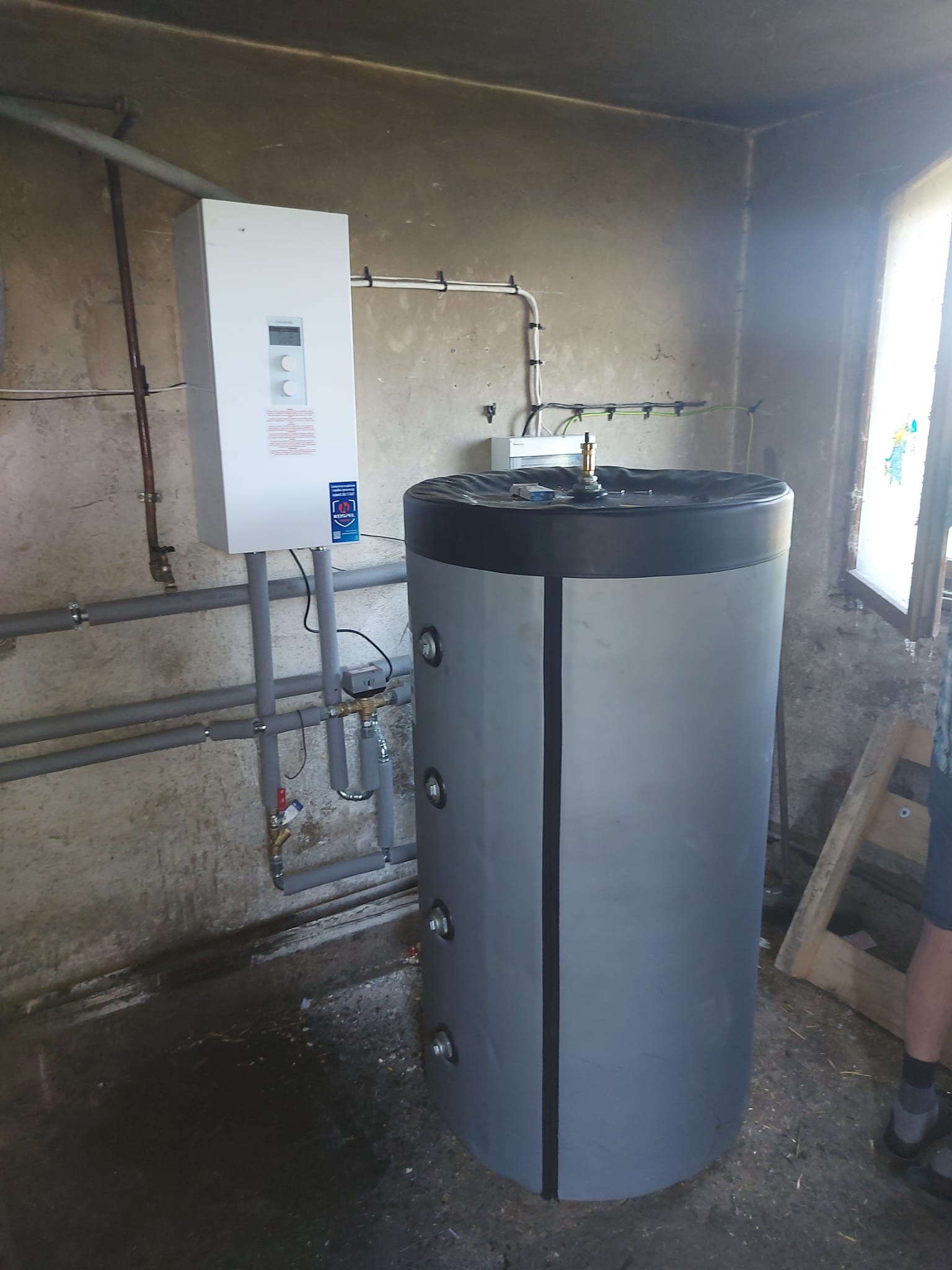 Szara, cylindryczna obudowa zbiornika buforowego z czarną górną pokrywą, połączona rurami z białym, prostokątnym panelem sterującym na tle surowej ściany w pomieszczeniu gospodarczym.