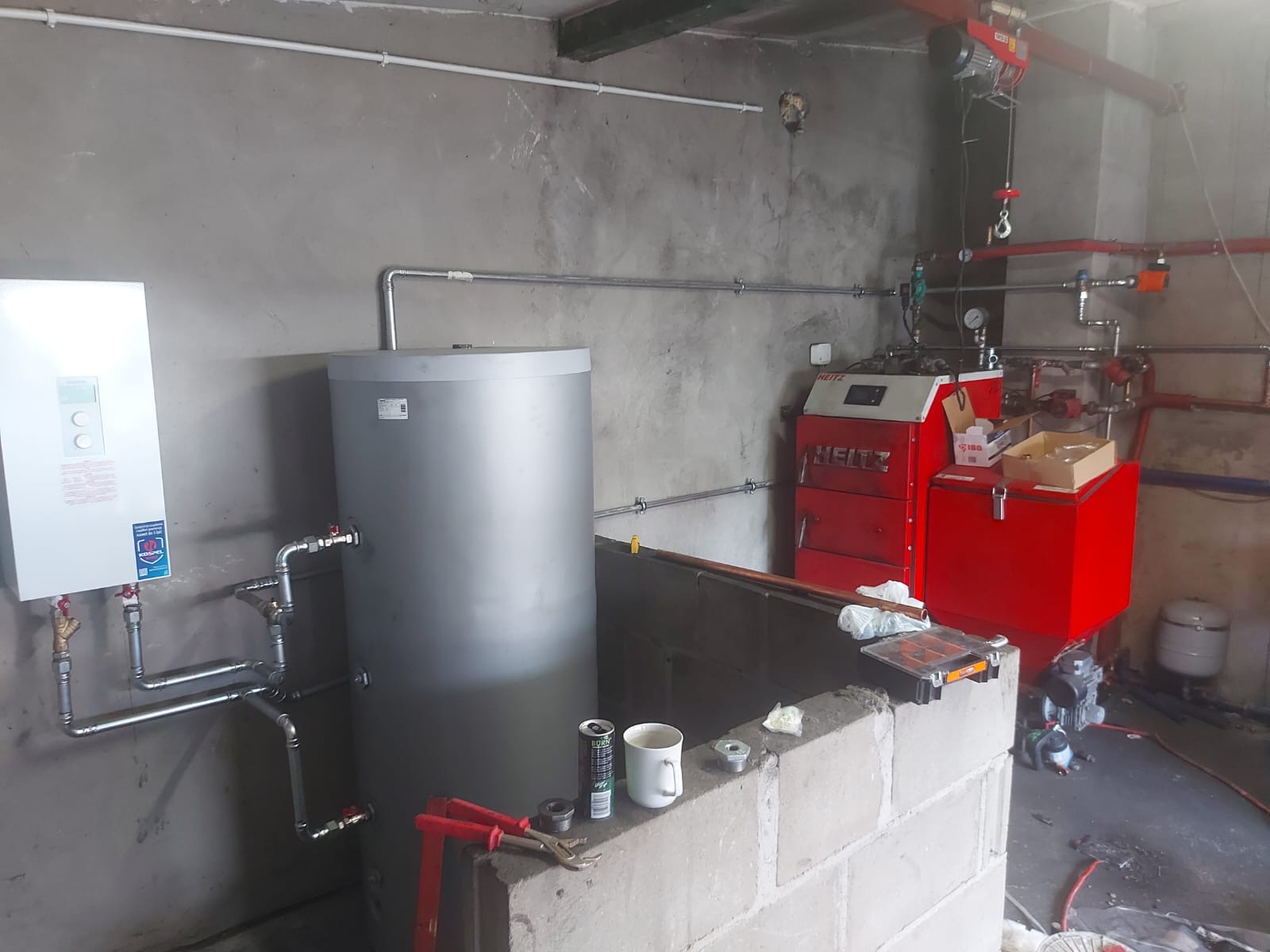 Szary zbiornik buforowy i biały moduł pompy ciepła zamontowane w surowym pomieszczeniu technicznym z betonowymi ścianami, widoczny również czerwony kocioł grzewczy Heitz, narzędzia i puszka napoju...